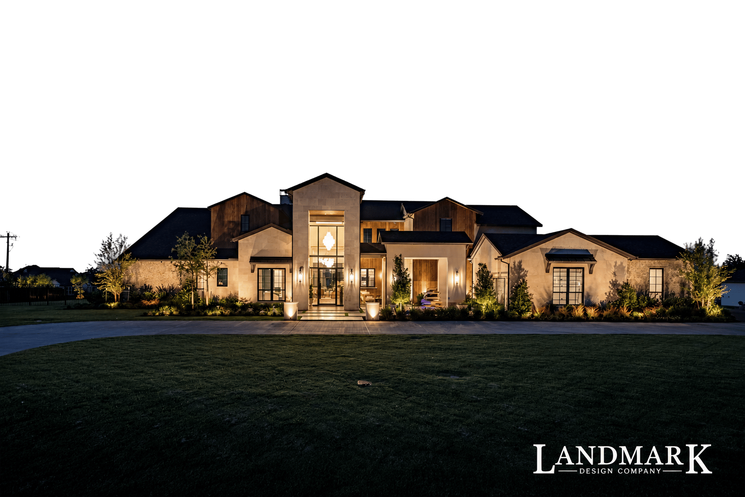 Stone Mediterranean-style home outlined in warm roofline lights, with uplit trees along a paver driveway at night