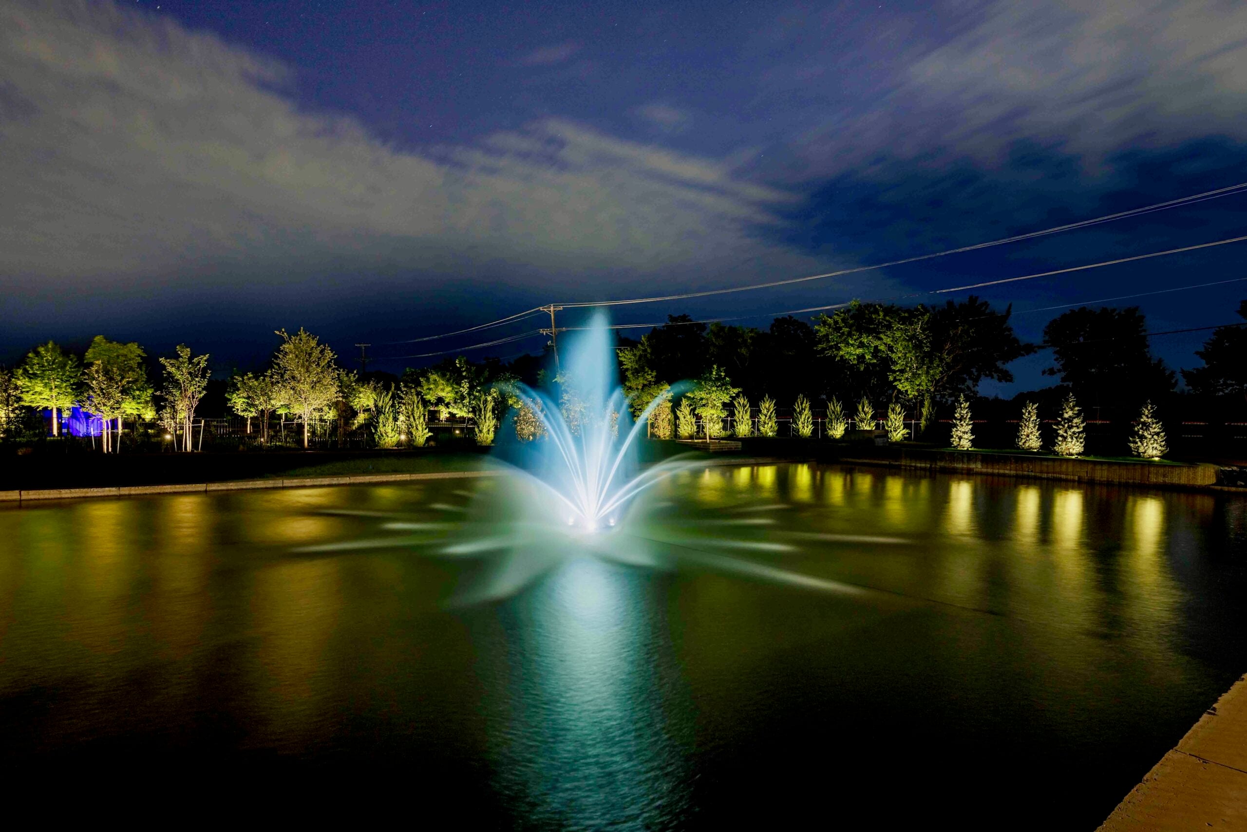 Illuminated fountain in a pond at night, surrounded by trees and reflecting lights on the water surface.