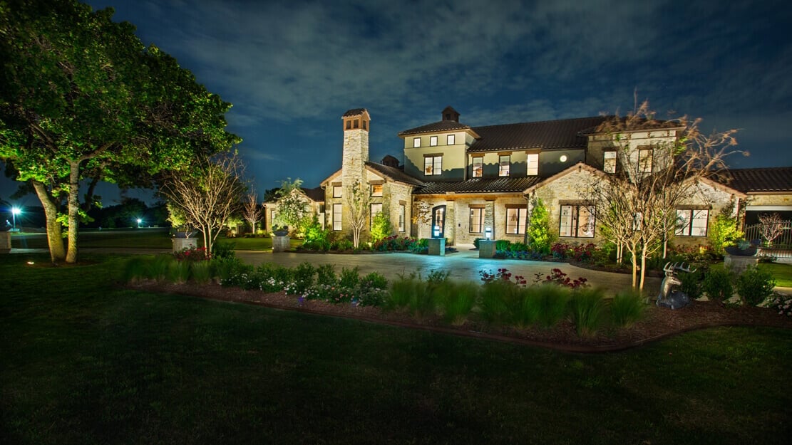 Illuminated stone house at night surrounded by trees and landscaped garden.