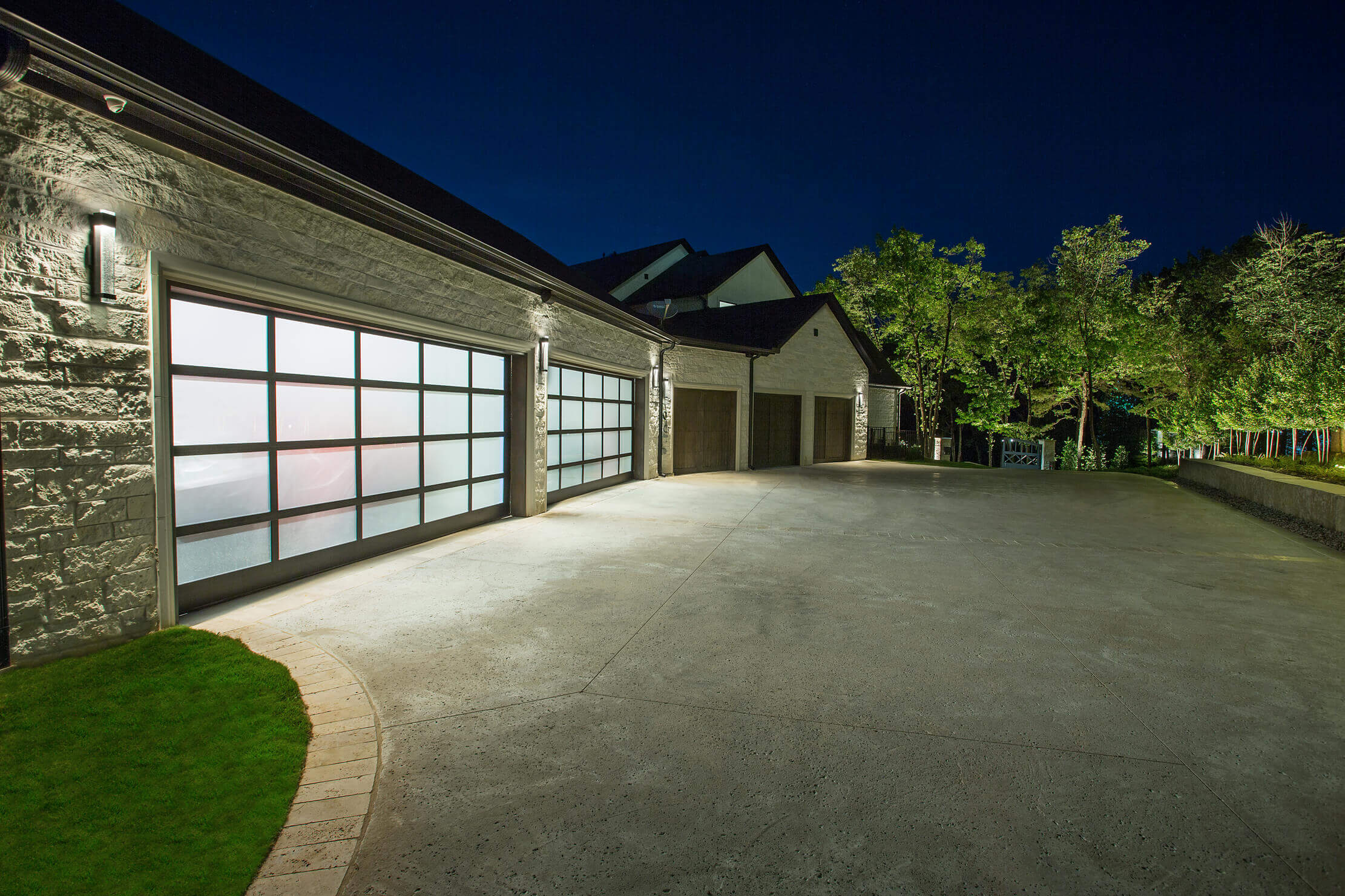 A modern garage with glass doors, illuminated at night, beside a concrete driveway and green grass.