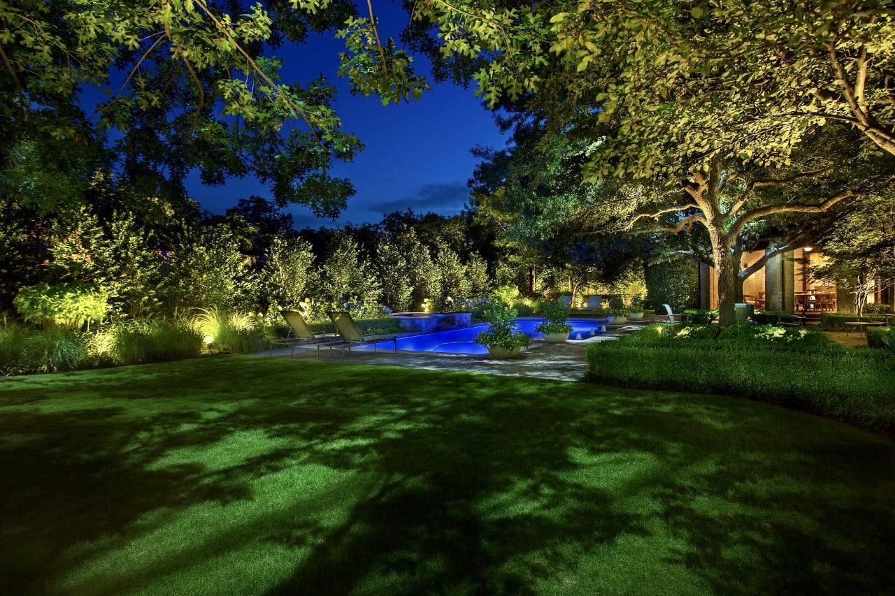 A beautifully lit backyard at night featuring a pool, lounge chairs, and lush greenery under tall trees.