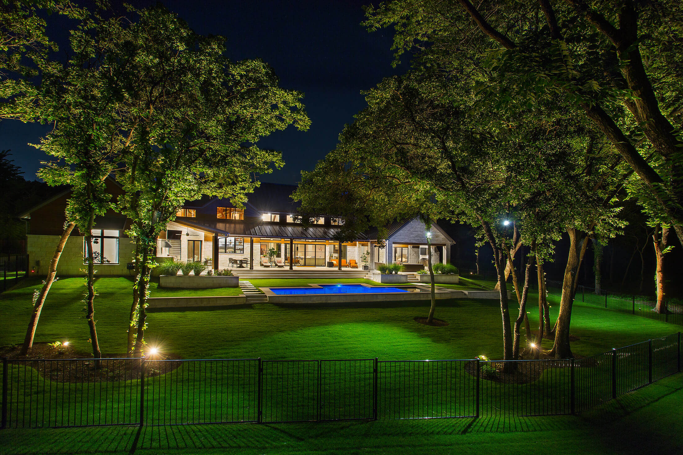 Modern house with large windows and a pool, surrounded by trees and well-lit garden, at night.
