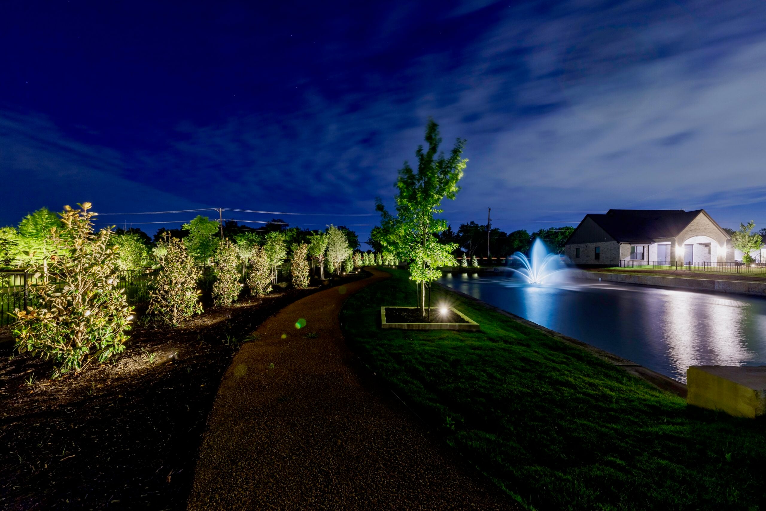A nighttime scene featuring a path beside a lit pond and fountain, bordered by illuminated trees and a house.