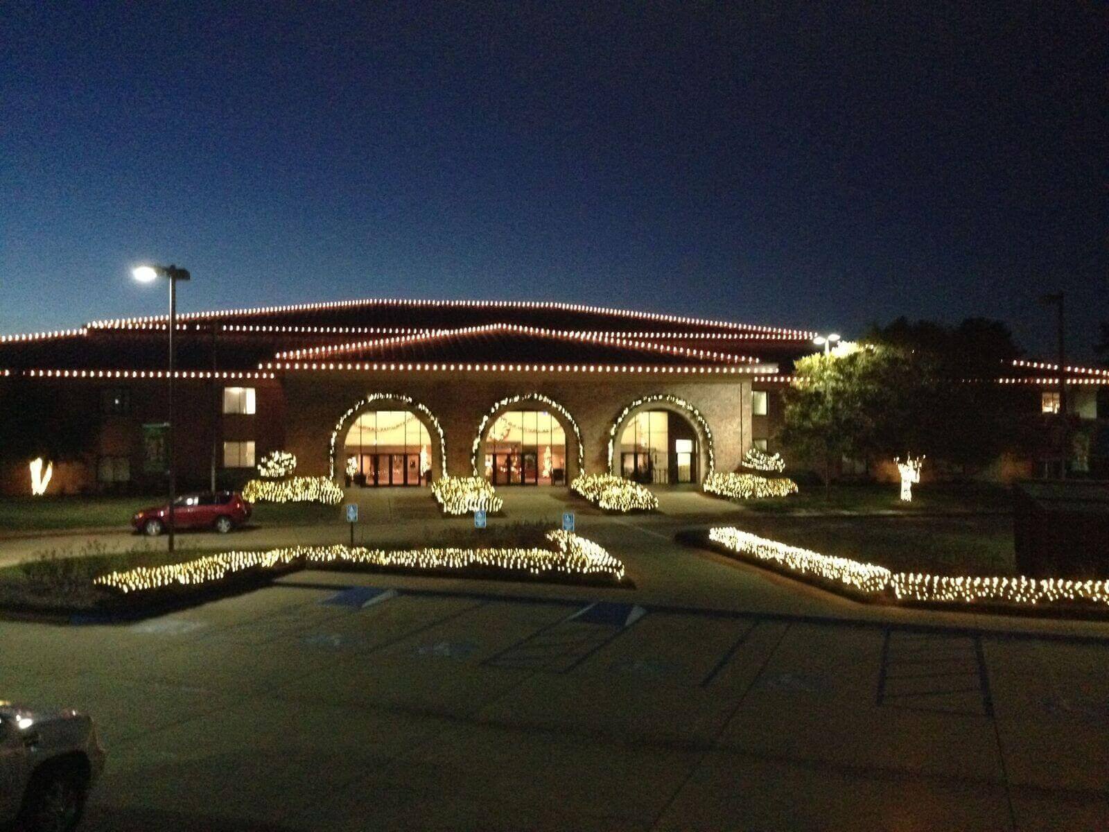 A brick building decorated with white lights at night, surrounded by shrubs and trees lit with holiday lights.