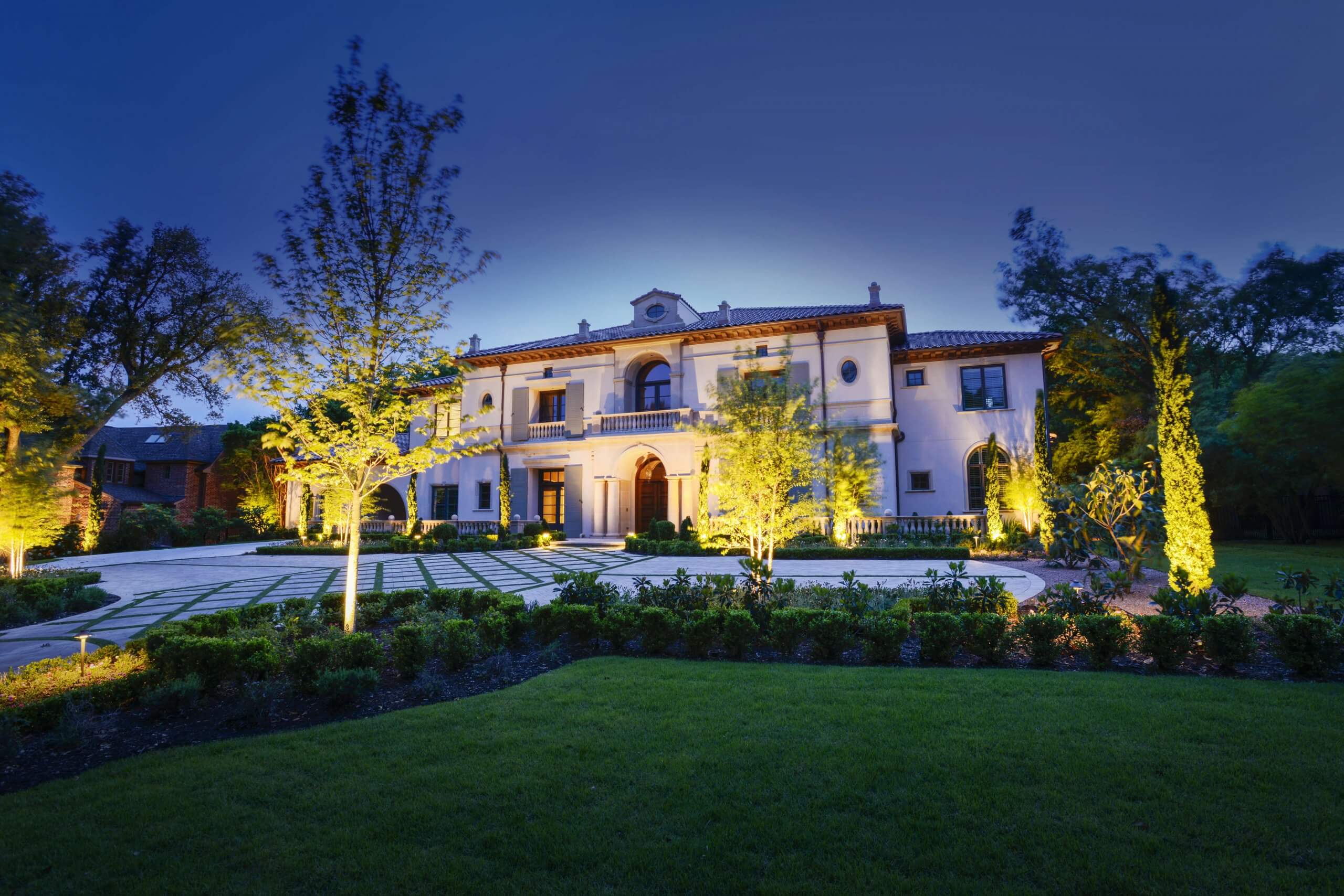 A villa façade and paved front courtyard featuring bright landscape spotlighting across surrounding young trees and lush lawn spaces.