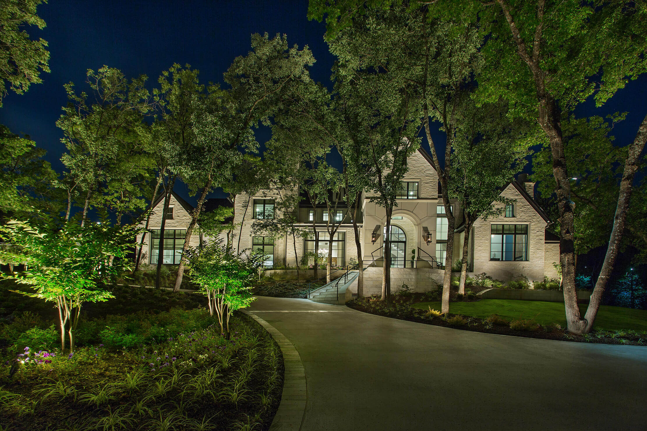 A modern home illuminated at night, surrounded by trees and landscaped gardens, with a walkway leading to the entrance.