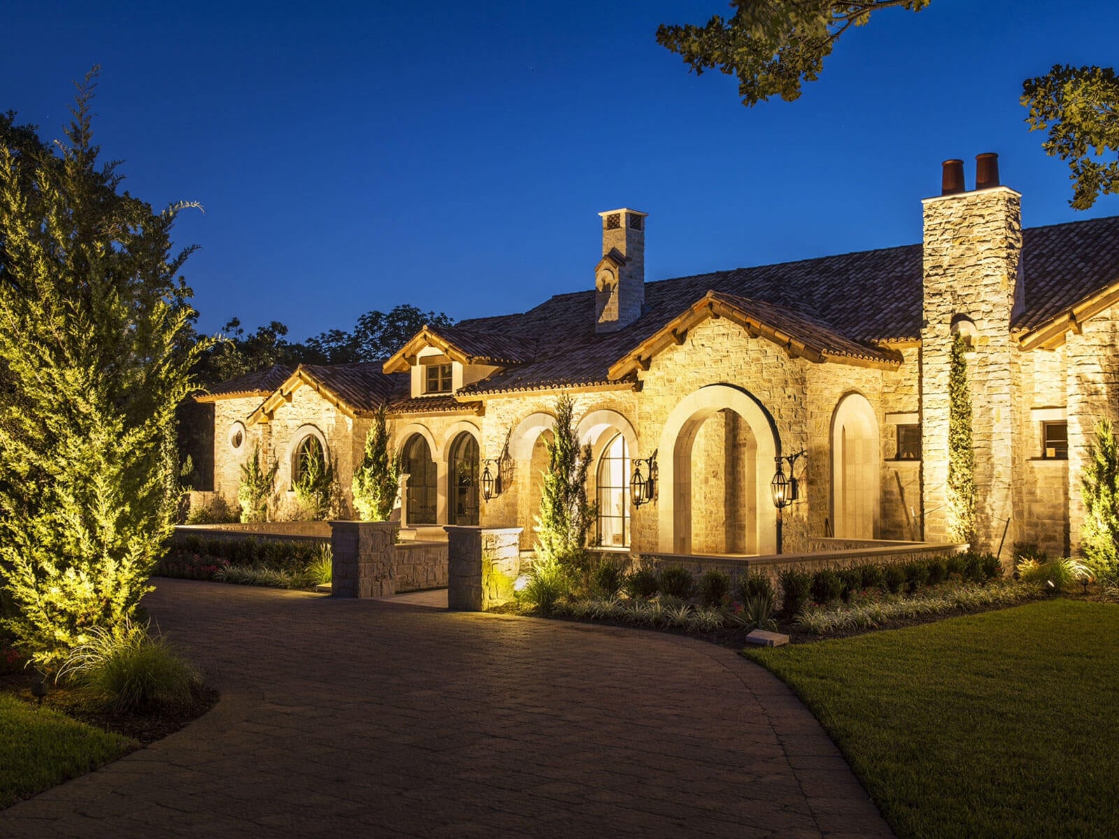 A close-up view of stone archways and entryways illuminated by warm exterior lighting alongside paved driveway landscaping.
