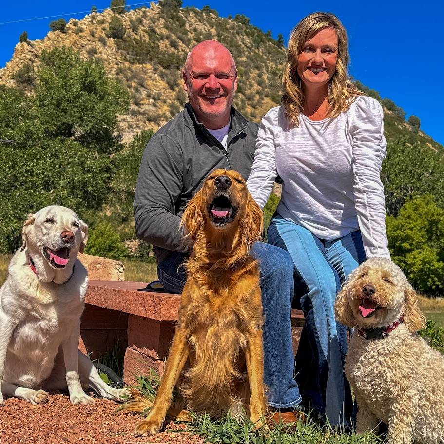 A man and a woman sit on a bench with three dogs, smiling outdoors with mountains and blue sky in the background.