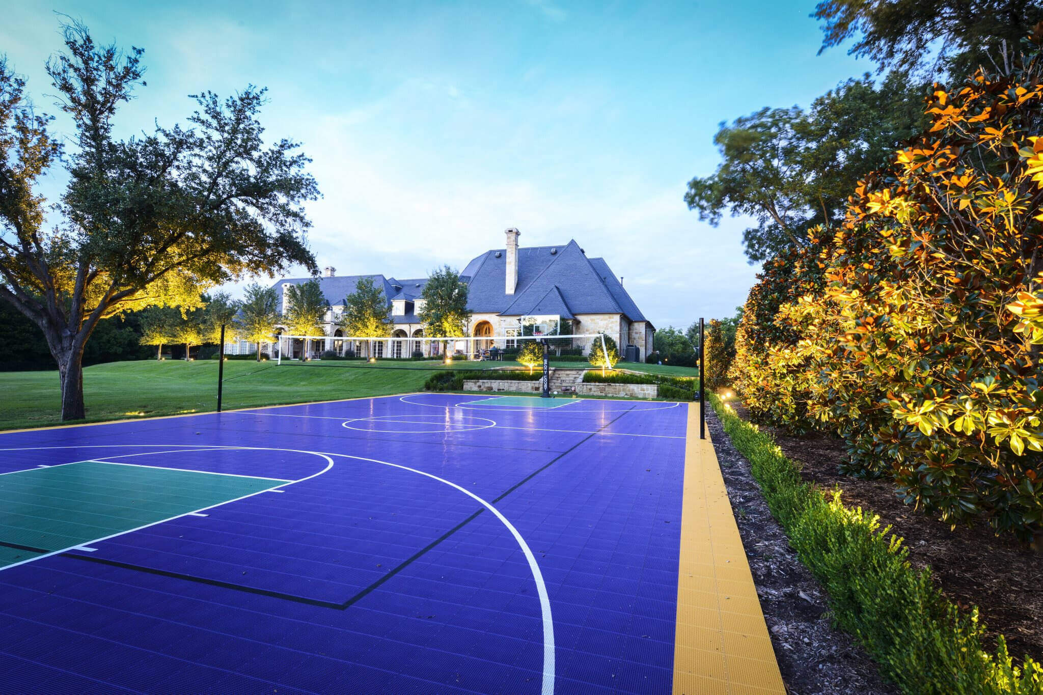 A colorful blue and green sports court set beside a lawn with landscape lighting casting a warm glow on surrounding trees and the home in the background.