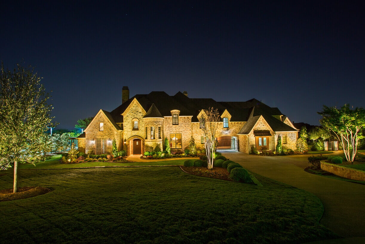 A wide night view of a large stone estate showcasing warm architectural illumination across the building and surrounding trees.