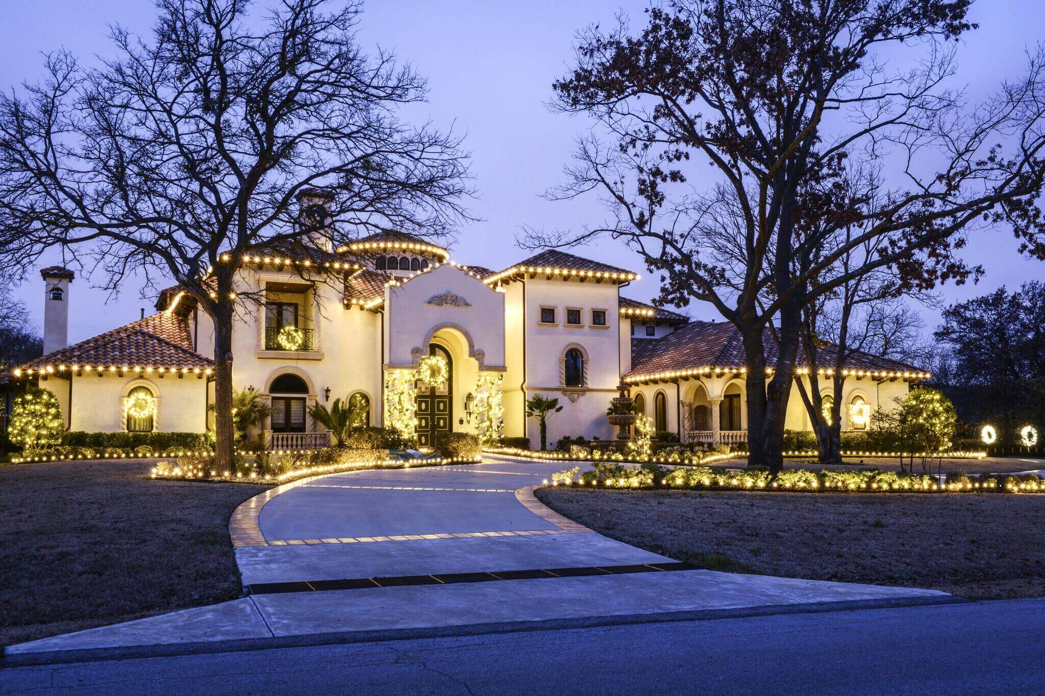 A beautifully lit, large white villa with a red tiled roof, surrounded by trees and festive lights at dusk.
