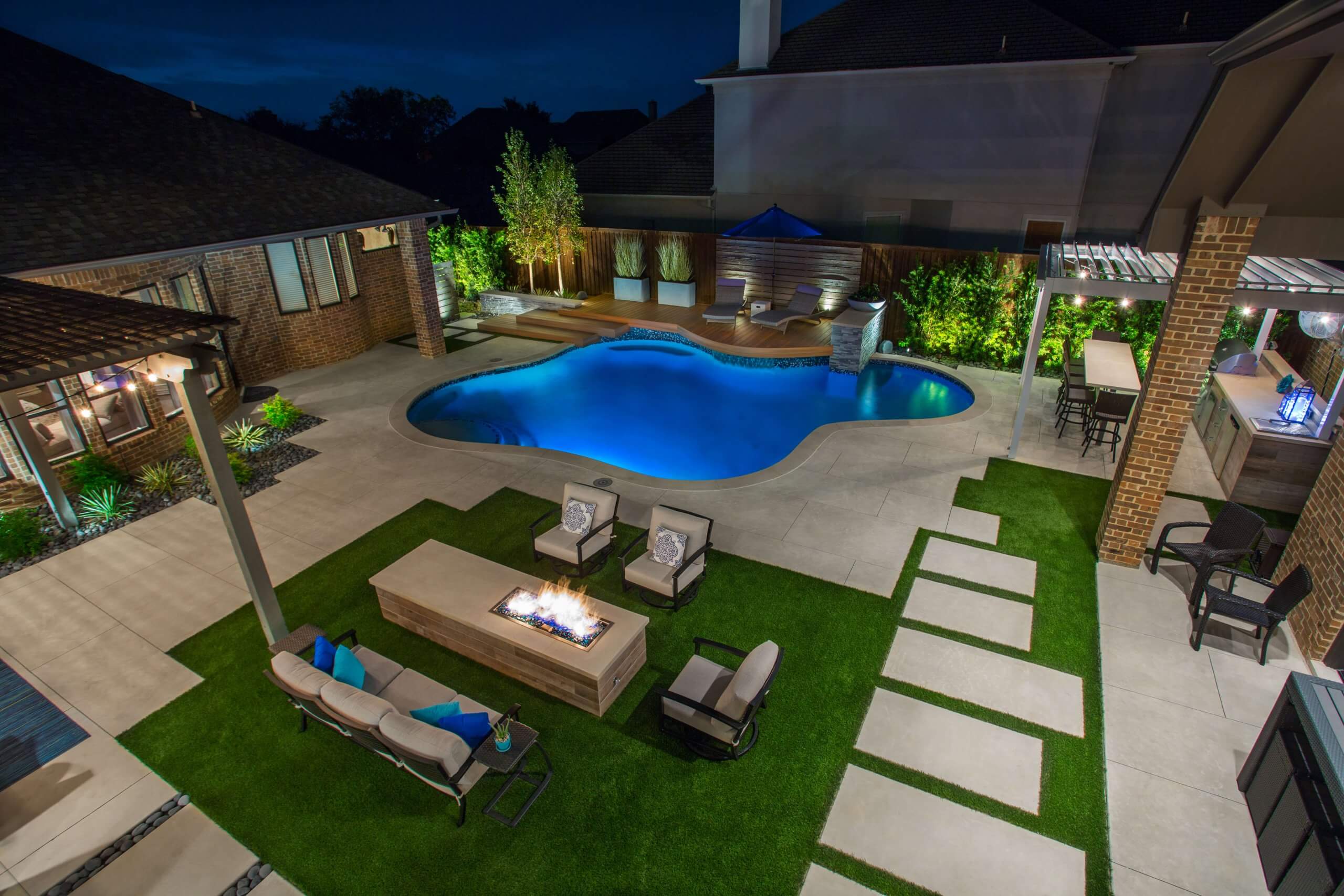 An overhead view of a luxury pool area and lawn patio featuring glowing pool lights, a modern gas fire pit, and uplit green foliage.