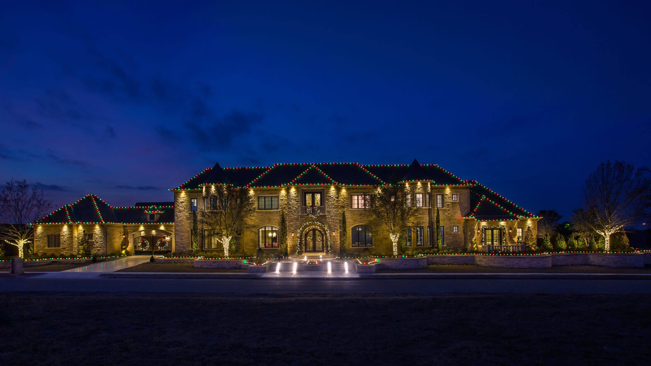 A large brick house decorated with colorful holiday lights at dusk, featuring trees and a walkway.