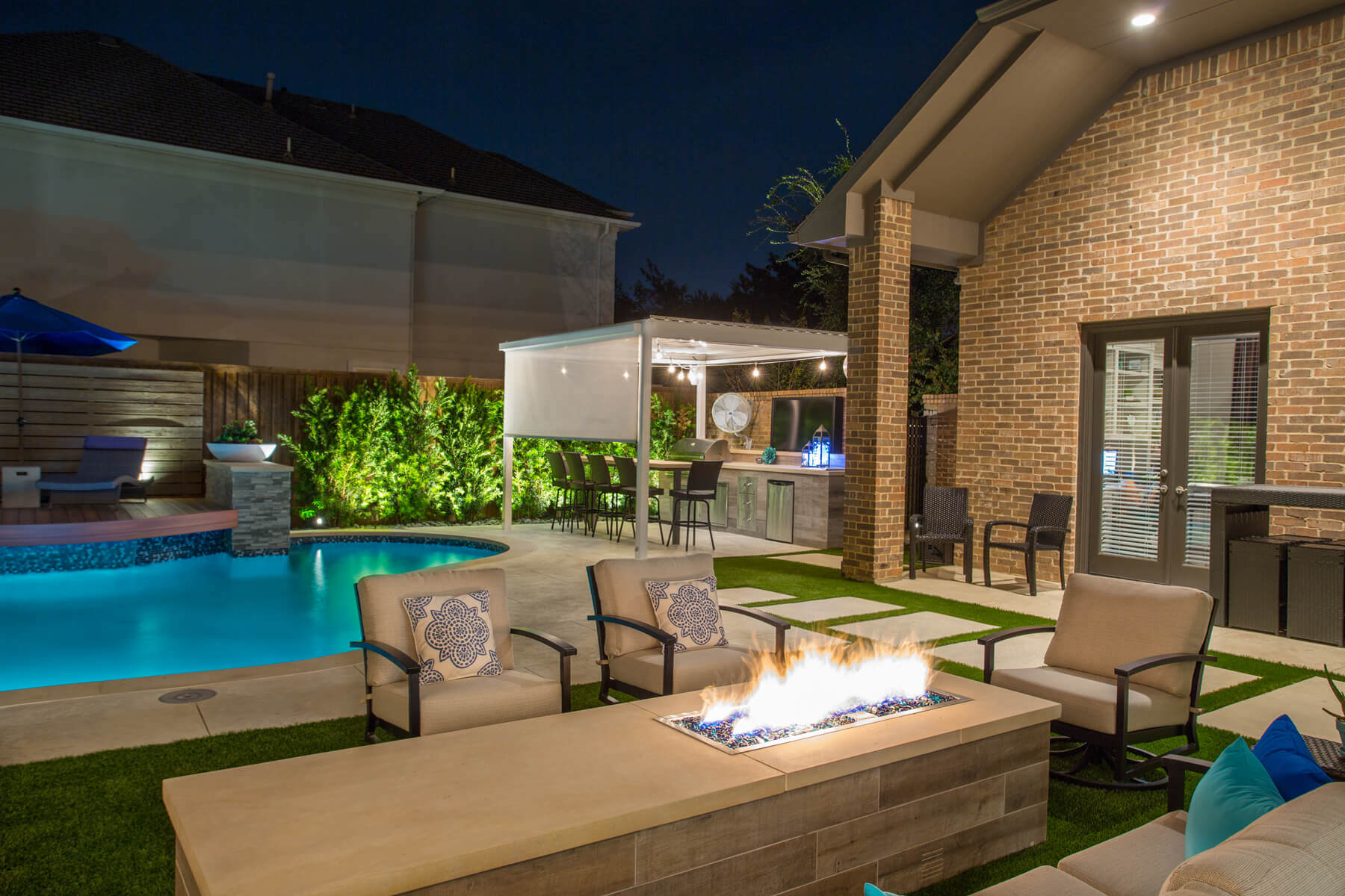 A close-up view of an outdoor lounge area centered around a lit gas fire pit beside a blue illuminated swimming pool and covered patio.