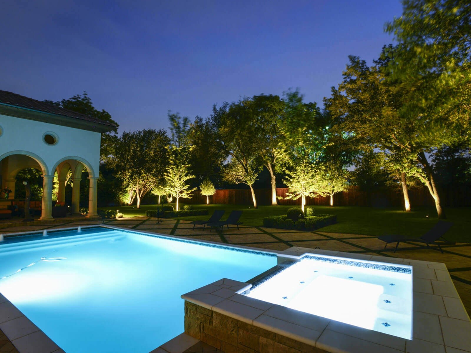 A bright blue pool and hot tub spa surrounded by a paver patio and mature lawn trees highlighted with warm accent uplighting.