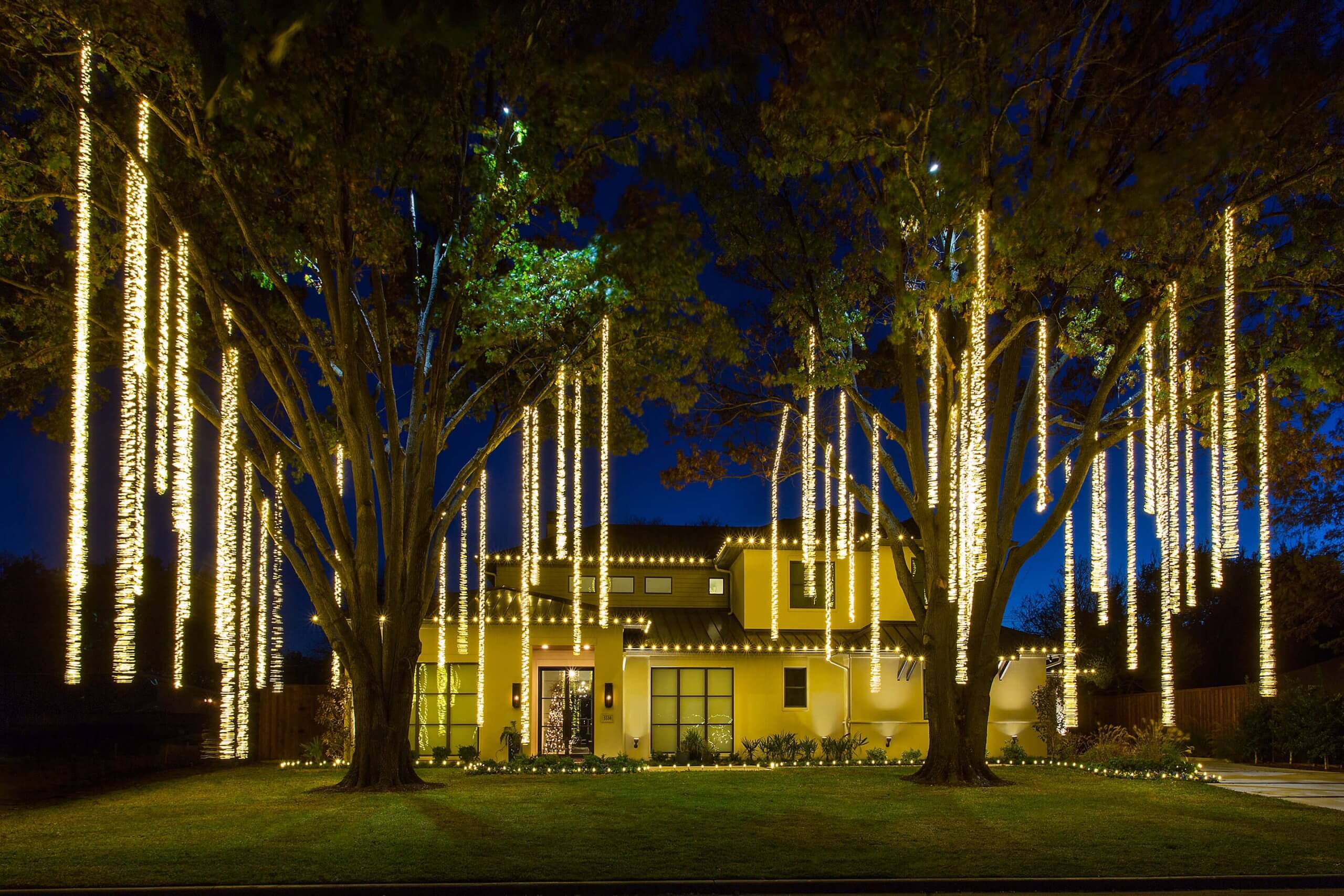A house at night adorned with bright, vertical string lights hanging from trees and along the roofline.
