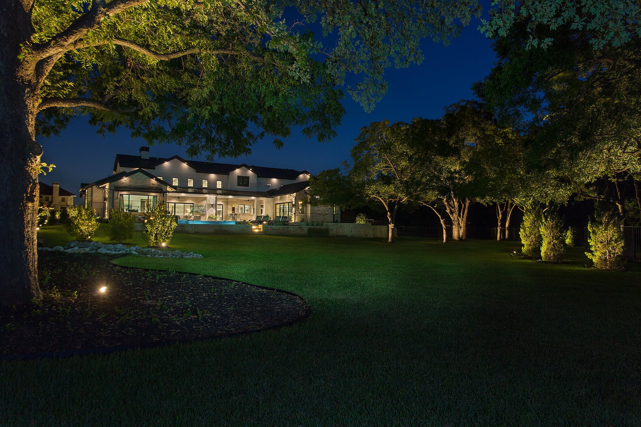 A illuminated modern house at night, surrounded by green lawn and trees, with outdoor lighting accentuating the landscaping.
