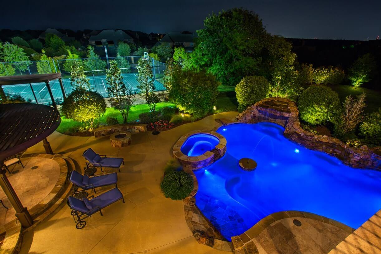 An elevated night view of a resort-style swimming pool glowing with vibrant blue lighting alongside uplit tropical trees.