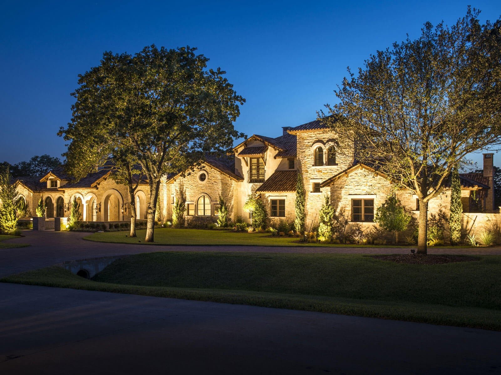 A sweeping view of a luxury residence driveway with warm lighting highlighting mature trees, stone walls, and arched entryways.
