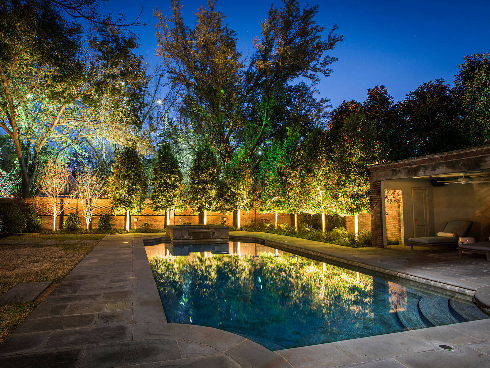 A rectangular pool reflecting ambient holiday light, bordered by stone pavers and a perimeter row of trees wrapped in warm landscape christmas lights.