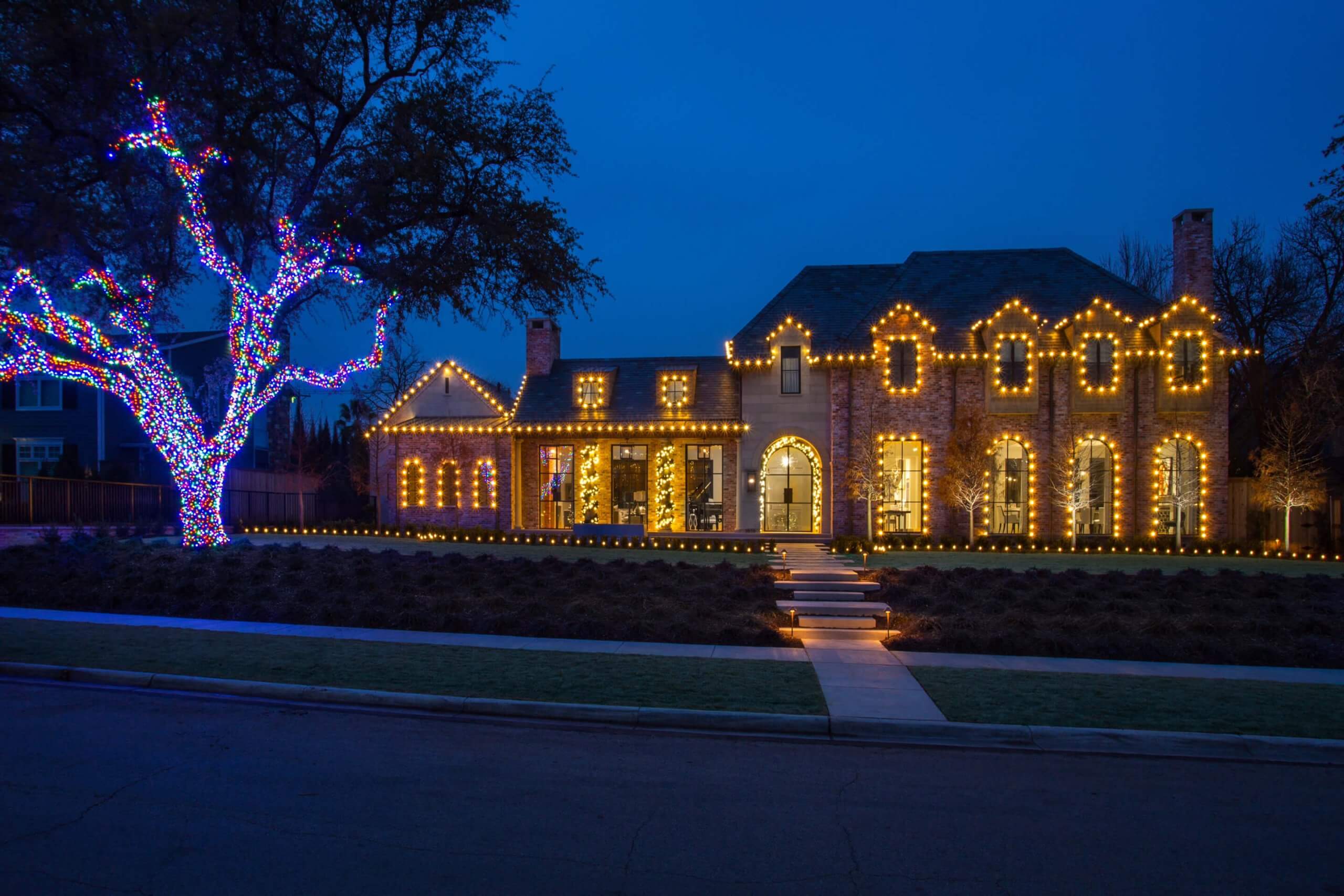 A festive house decorated with warm white and colorful Christmas lights at twilight, surrounded by a landscaped yard.