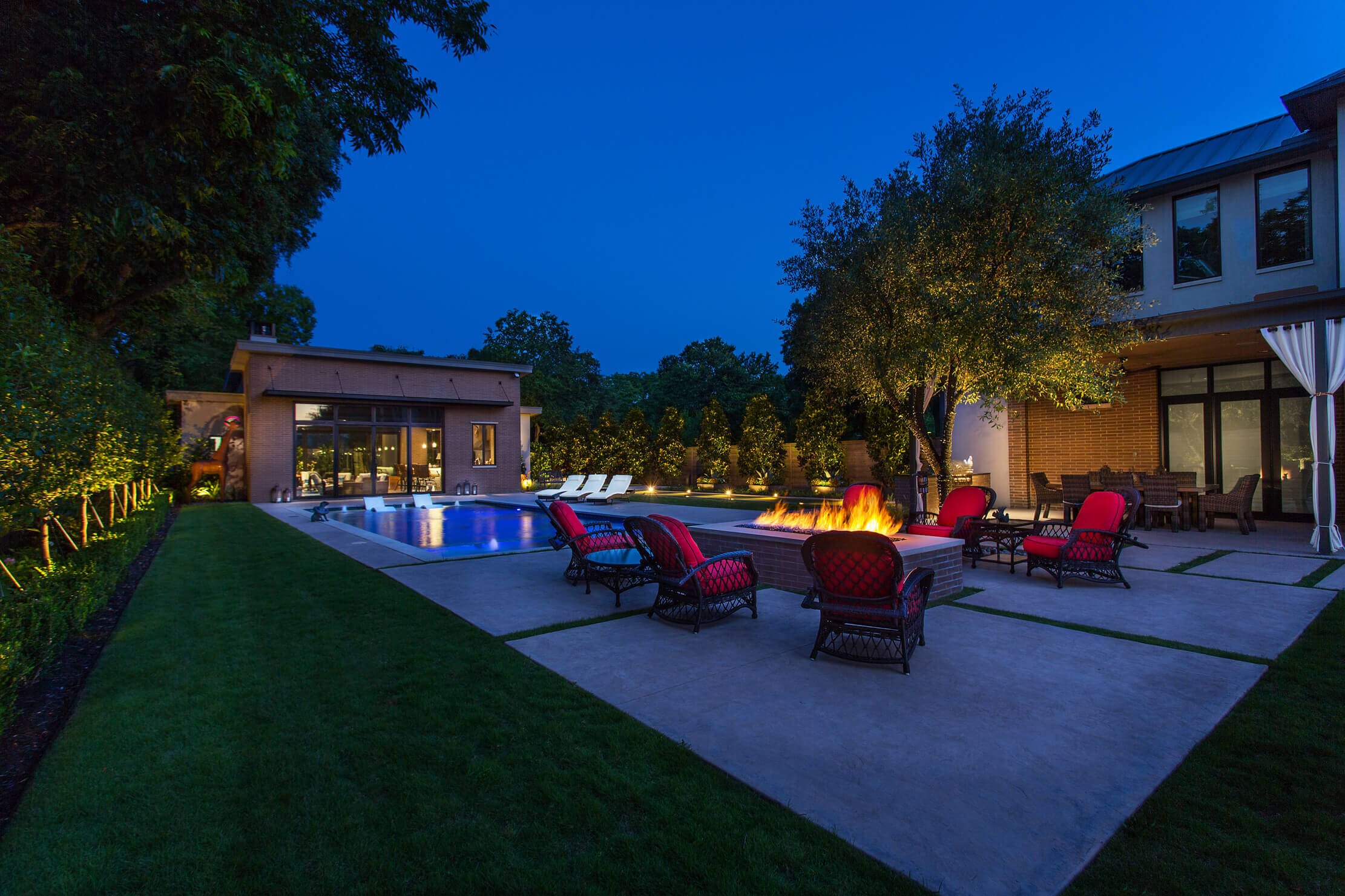 A modern backyard at night with a pool, fire pit, red lounge chairs, and decorative trees.