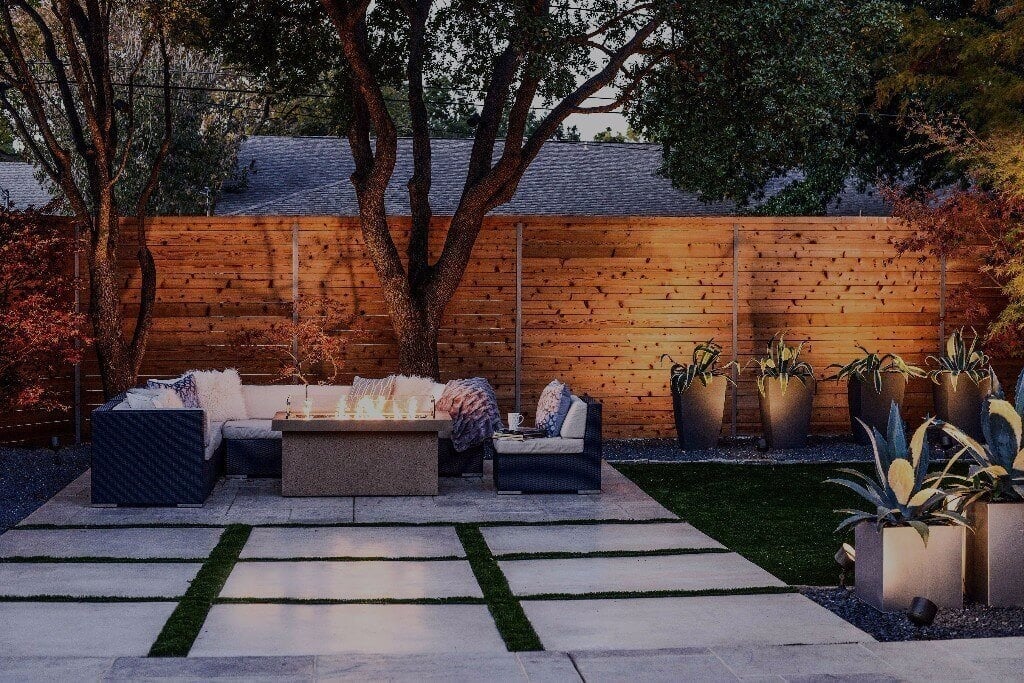 An outdoor seating lounge with a central fire table and warm ambient lighting accenting a wooden privacy fence and potted plants.