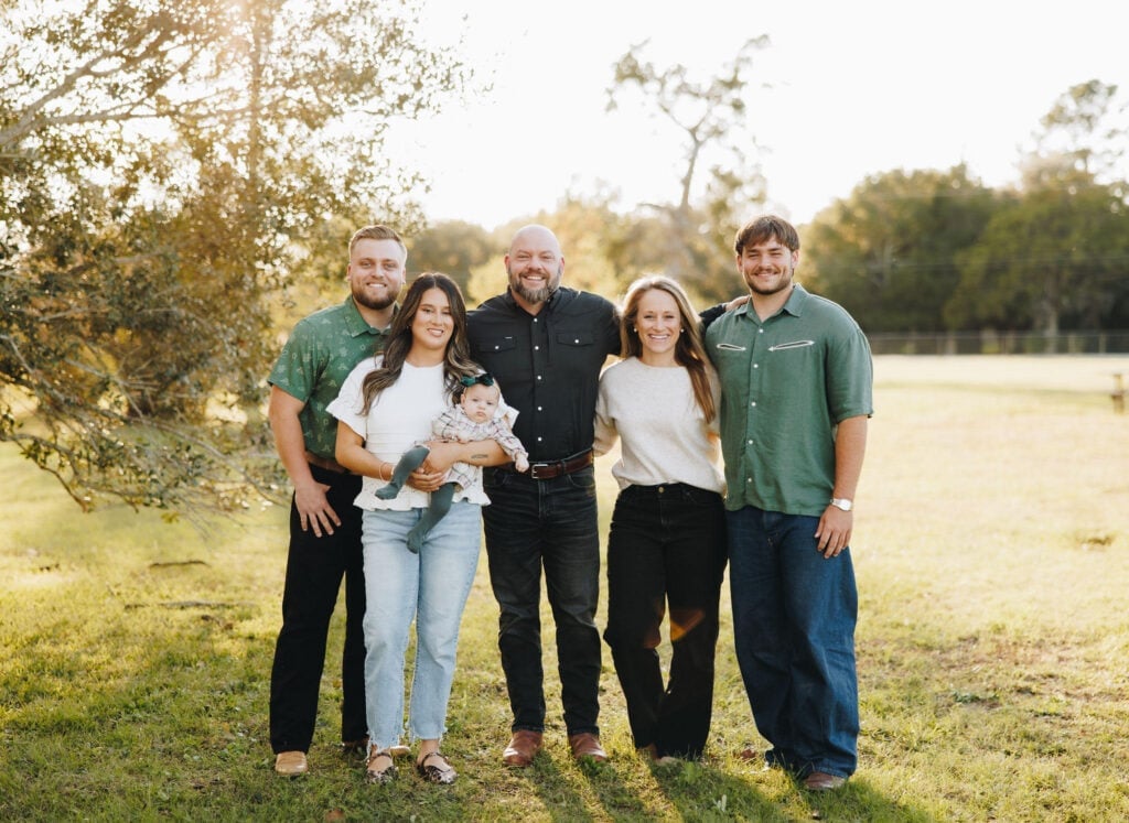Four people standing together outdoors, smiling in casual attire, with trees in the background.