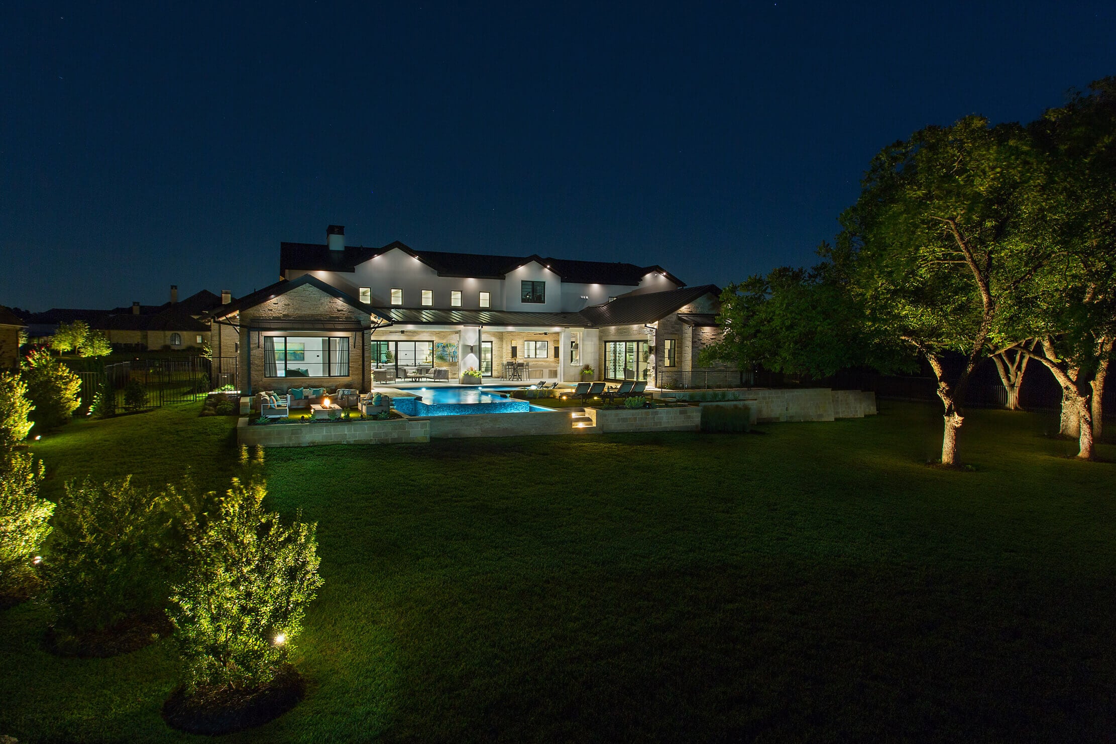 A modern house with large windows, pool, and well-lit yard at night, surrounded by trees and landscaping.
