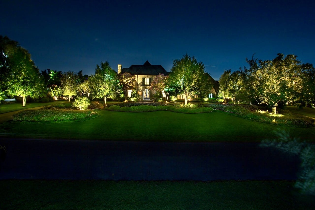 A stately home illuminated at night, surrounded by lush greenery and landscaped gardens.