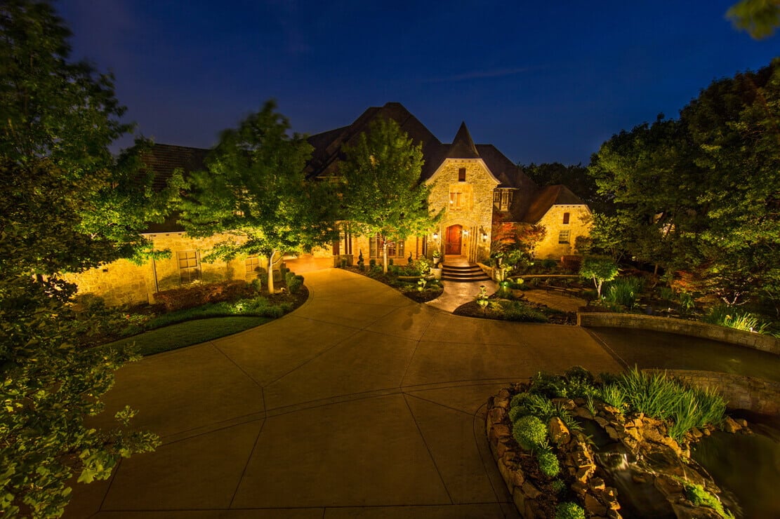 A paved driveway flanked by warm landscape uplighting on surrounding trees and stone home exterior.