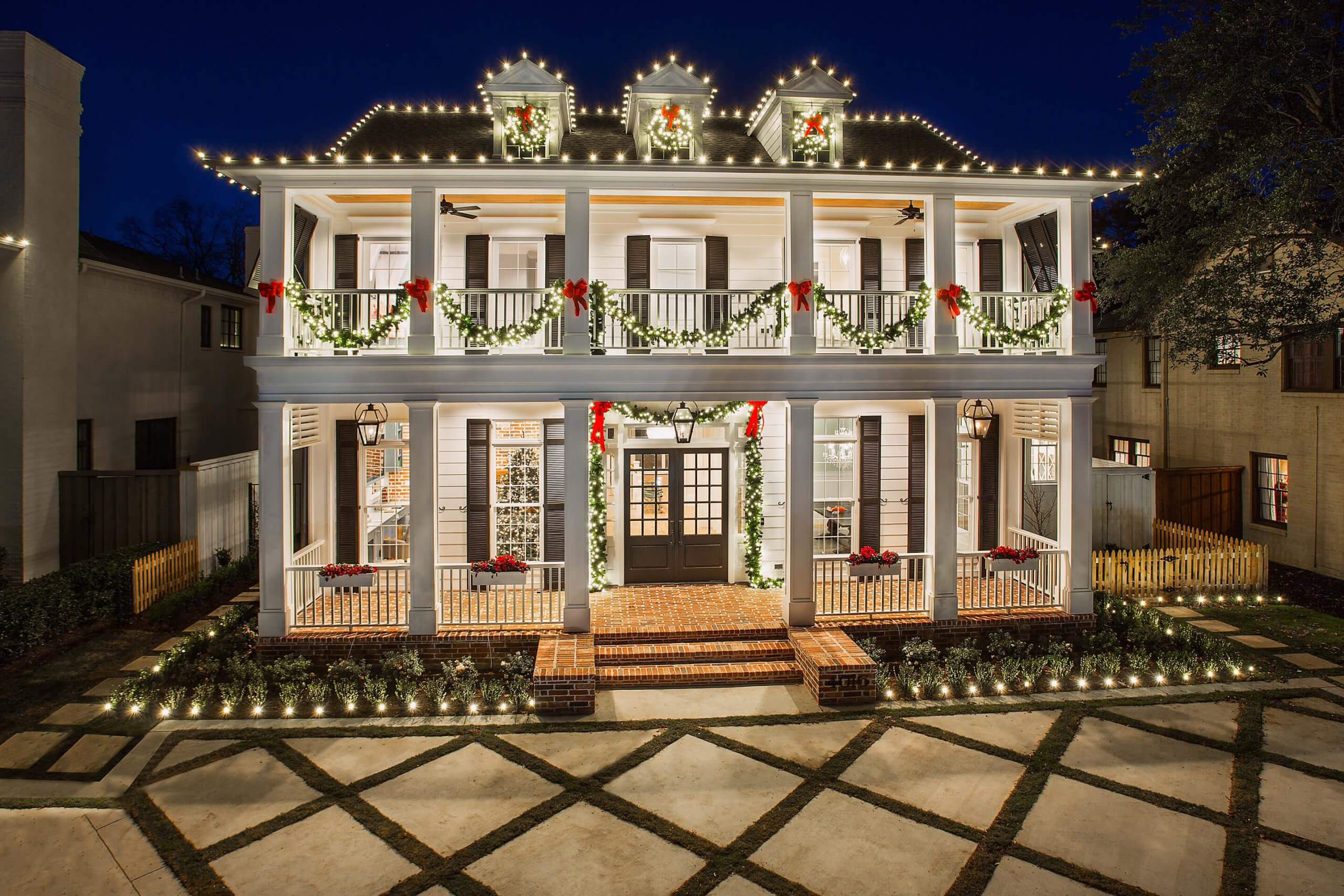 A beautifully decorated white house with holiday lights, garlands, and red bows, seen at night.