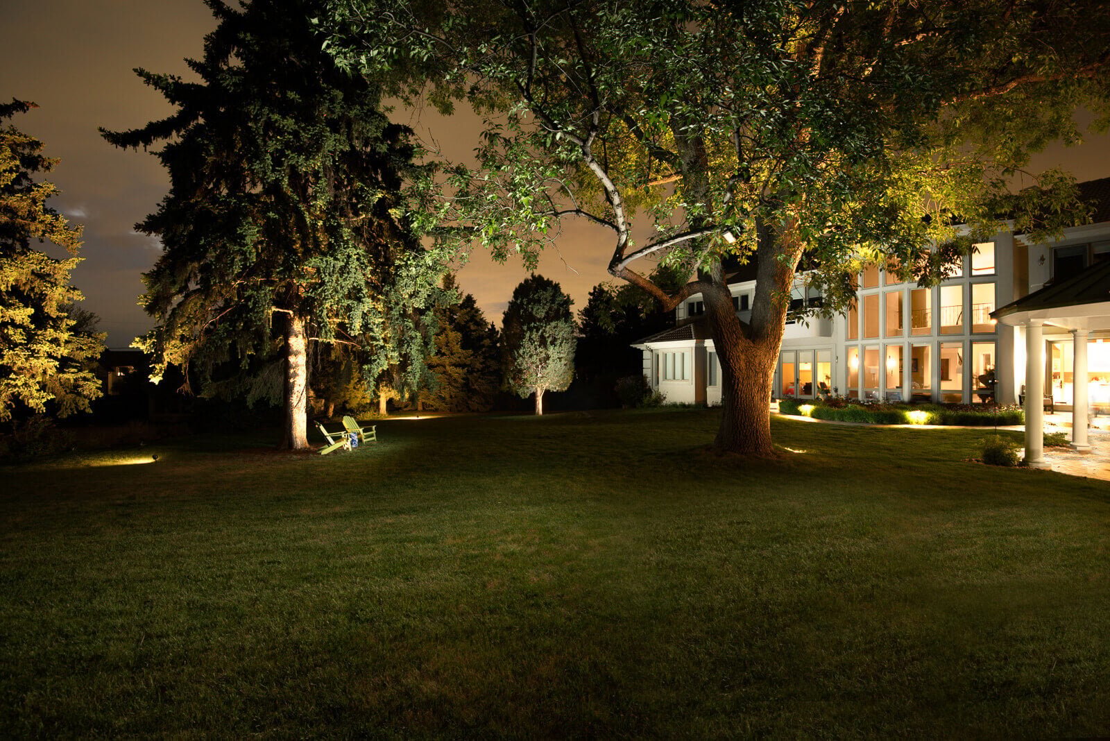 A night view of a large yard with trees, a house with illuminated windows, and a green chair in the grass.