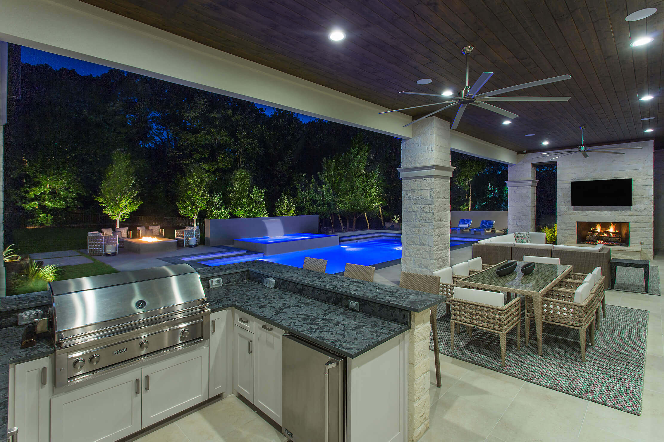 Luxurious outdoor patio with a grill, dining area, and pool, illuminated by soft lights at night.