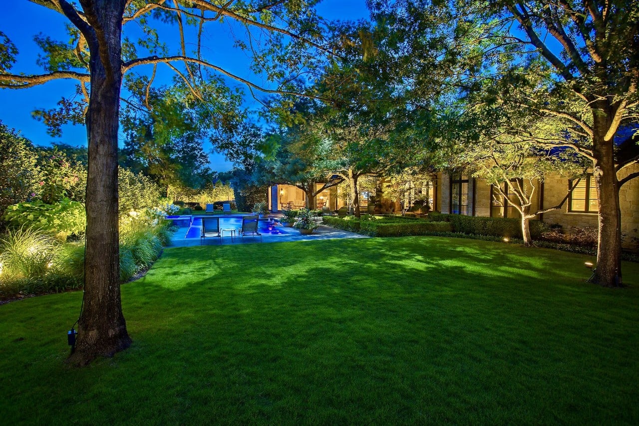 A well-lit backyard at dusk featuring a swimming pool, green lawn, and surrounding trees and shrubs.