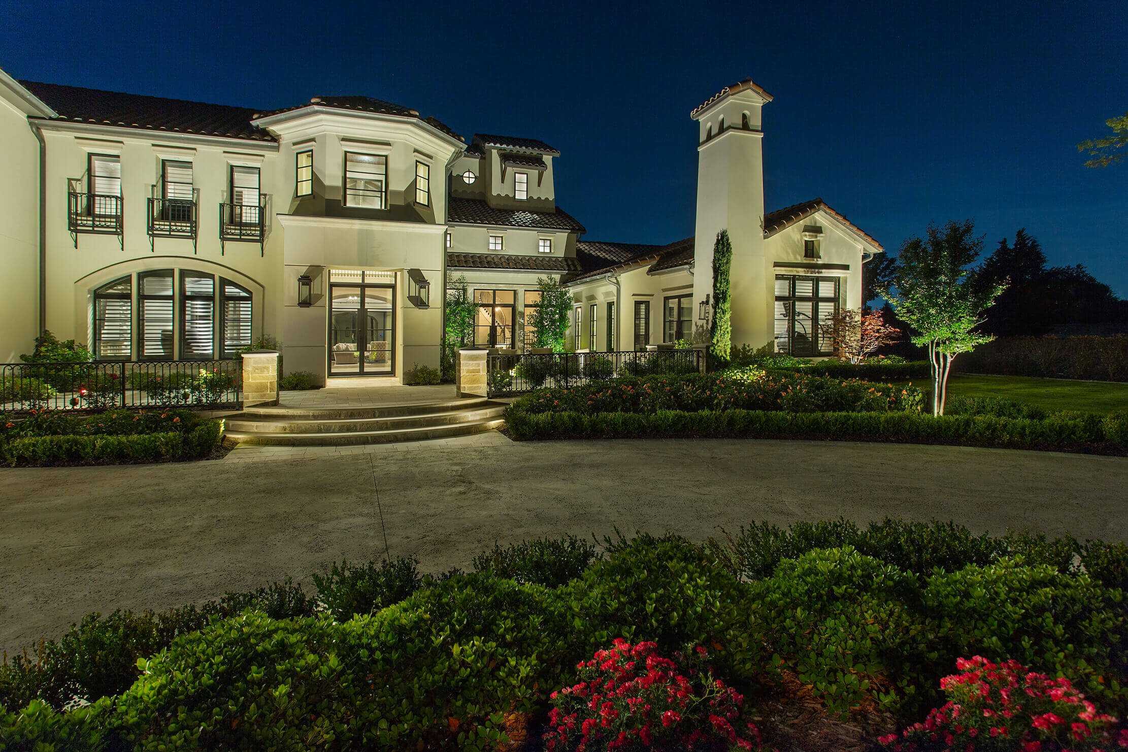 The front façade of a two-story luxury residence showcasing architectural accent lighting across stone columns, entryway steps, and manicured landscaping.