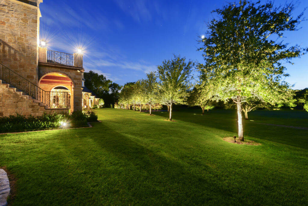 A spacious green lawn featuring a line of trees brightened by ground-level uplights next to a stone home exterior.