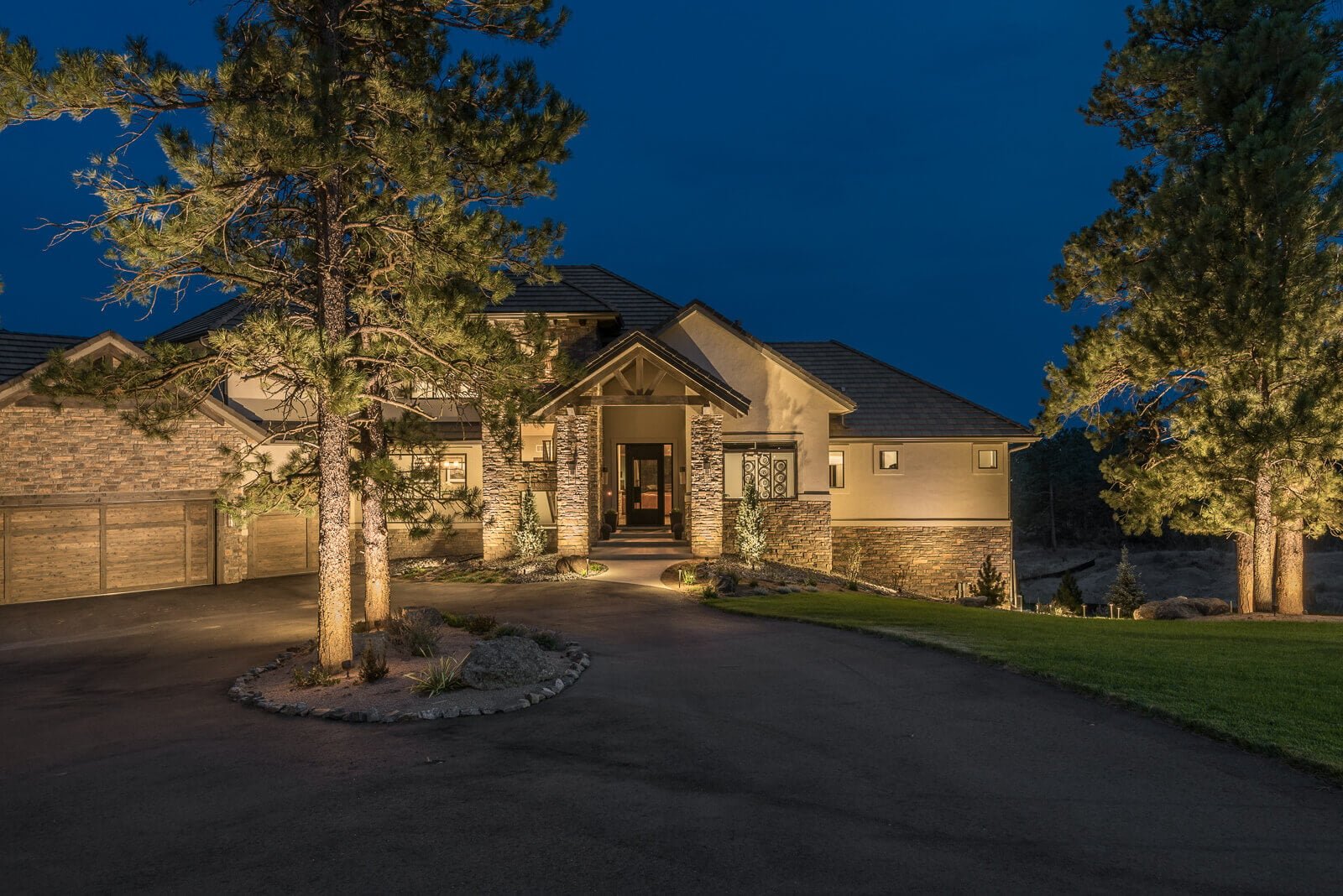 A modern house illuminated at night surrounded by trees and a driveway.