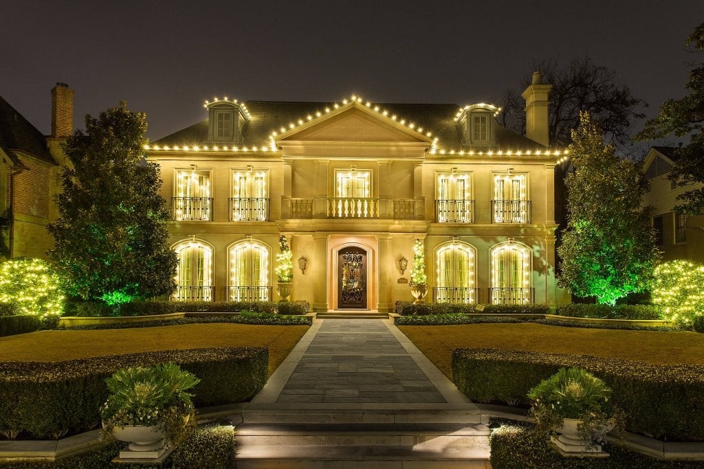 Two-story stone home with warm white roofline and window outlines, and green uplighting beside the entry steps.