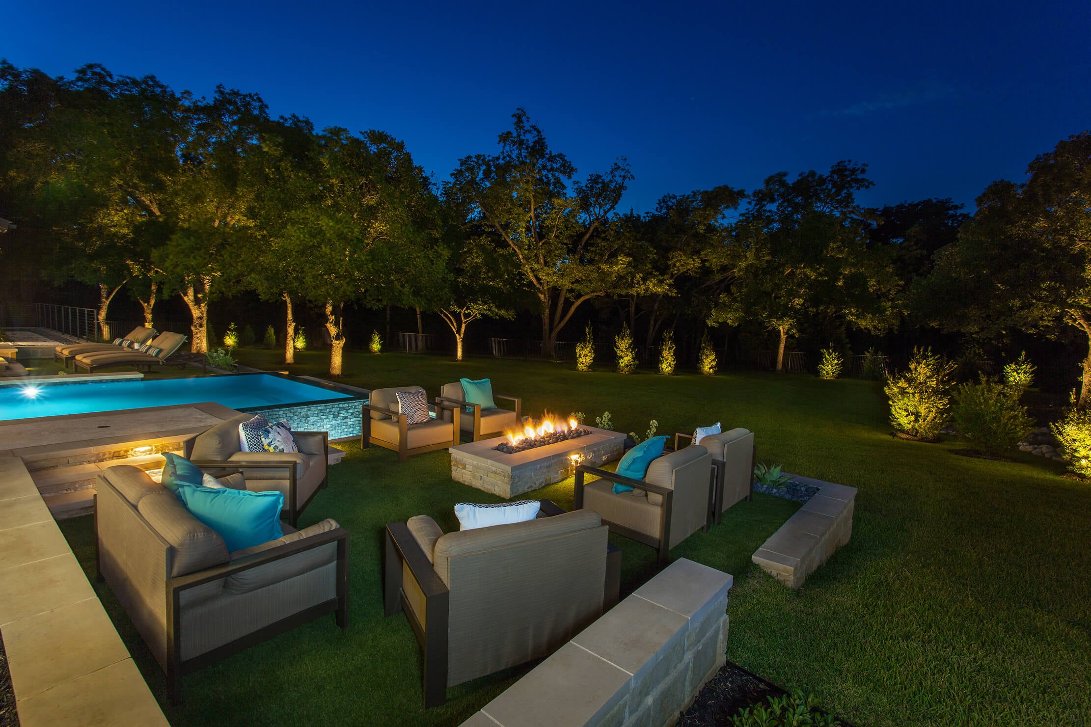 A backyard at night with seating around a fire pit, a pool, and trees illuminated by soft lighting.