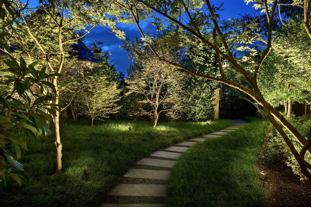 A serene garden pathway with stone tiles, surrounded by lush greenery and softly lit trees at dusk.