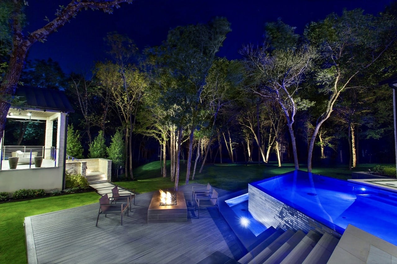 A modern backyard at night featuring a pool, fire pit, and landscaped trees under a starry sky.