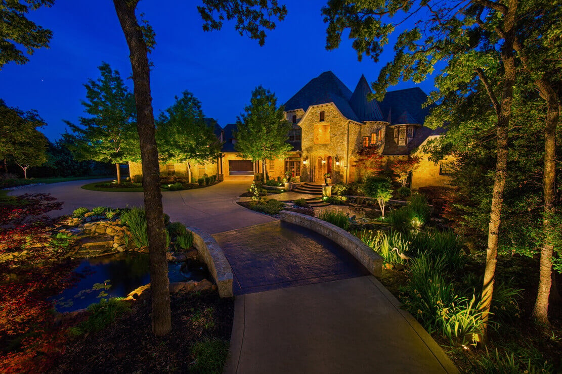A stone bridge entryway illuminated by warm landscape lighting, leading toward a stone home under a deep blue evening sky.