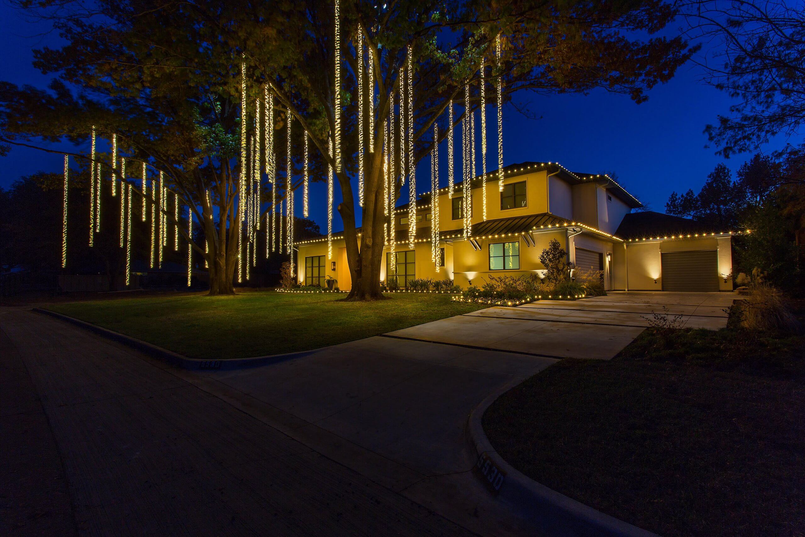 A well-lit house at night, decorated with hanging lights from trees and along the roof.