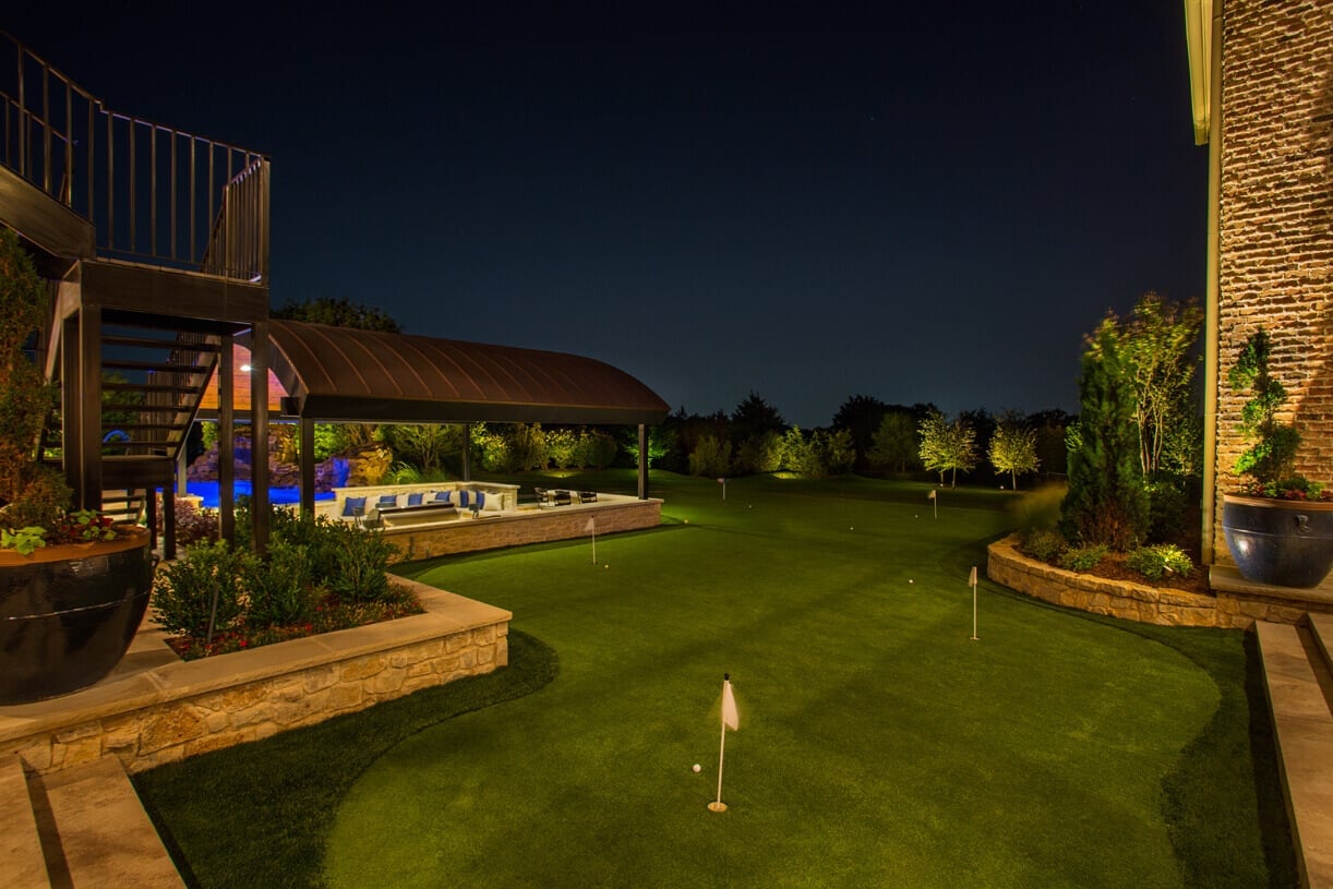 A private backyard putting green soft-lit at night with surrounding stone retaining walls and uplit garden foliage.