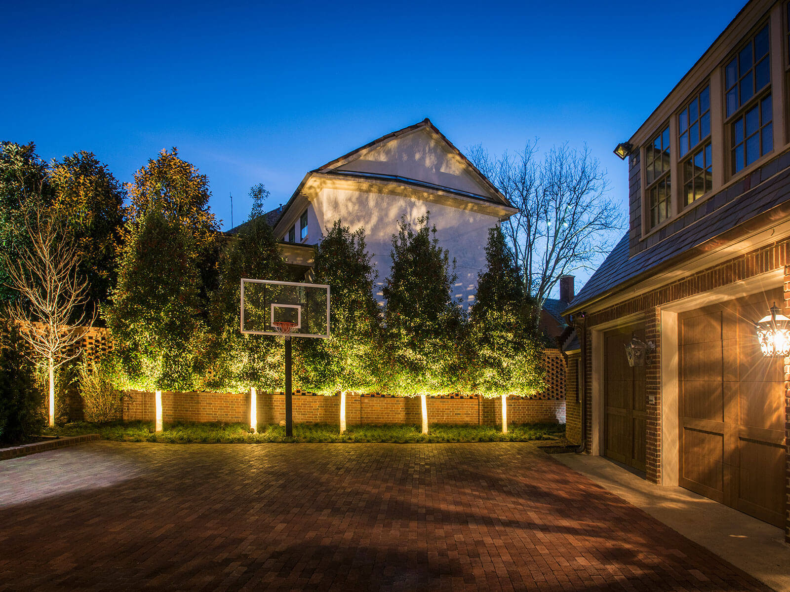 A paved backyard basketball court bordered by a dense line of privacy trees softly illuminated with festive base lighting along a brick fence line.