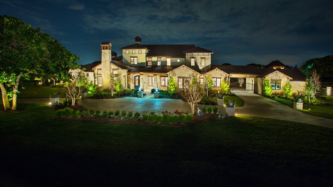 A beautifully illuminated two-story stone and stucco house at night, surrounded by landscaped gardens and trees.