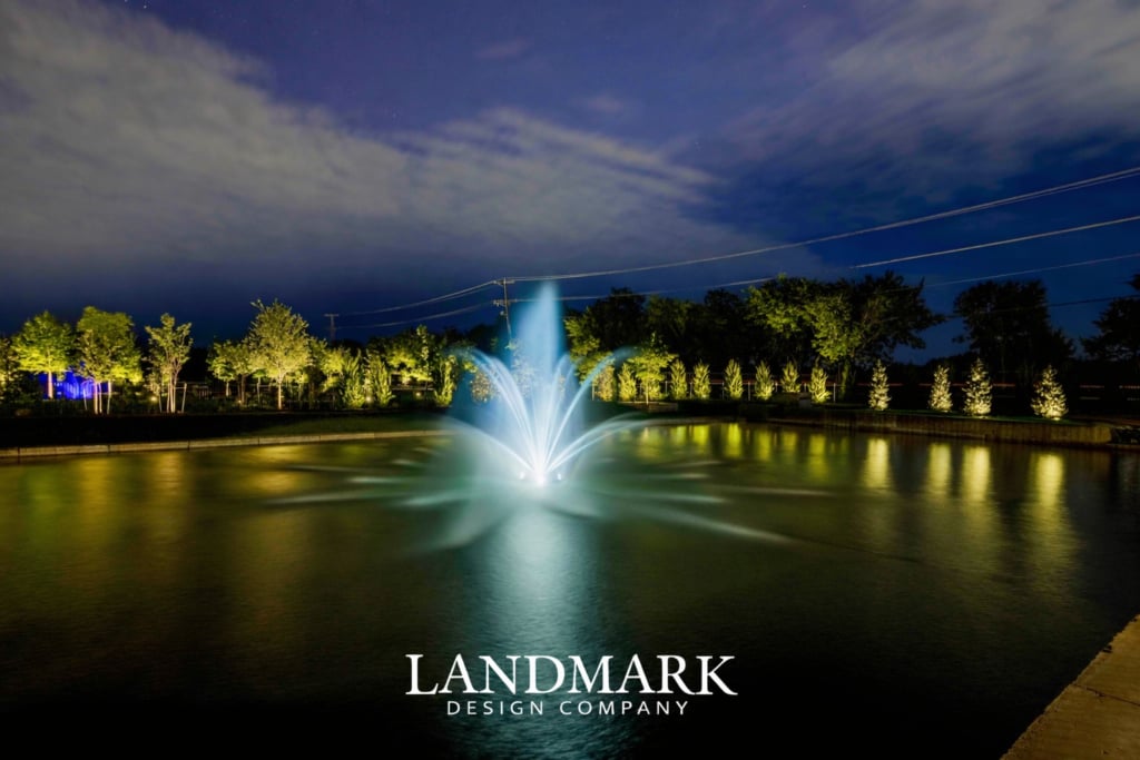 A illuminated fountain in a pond at night, surrounded by trees and soft lighting.