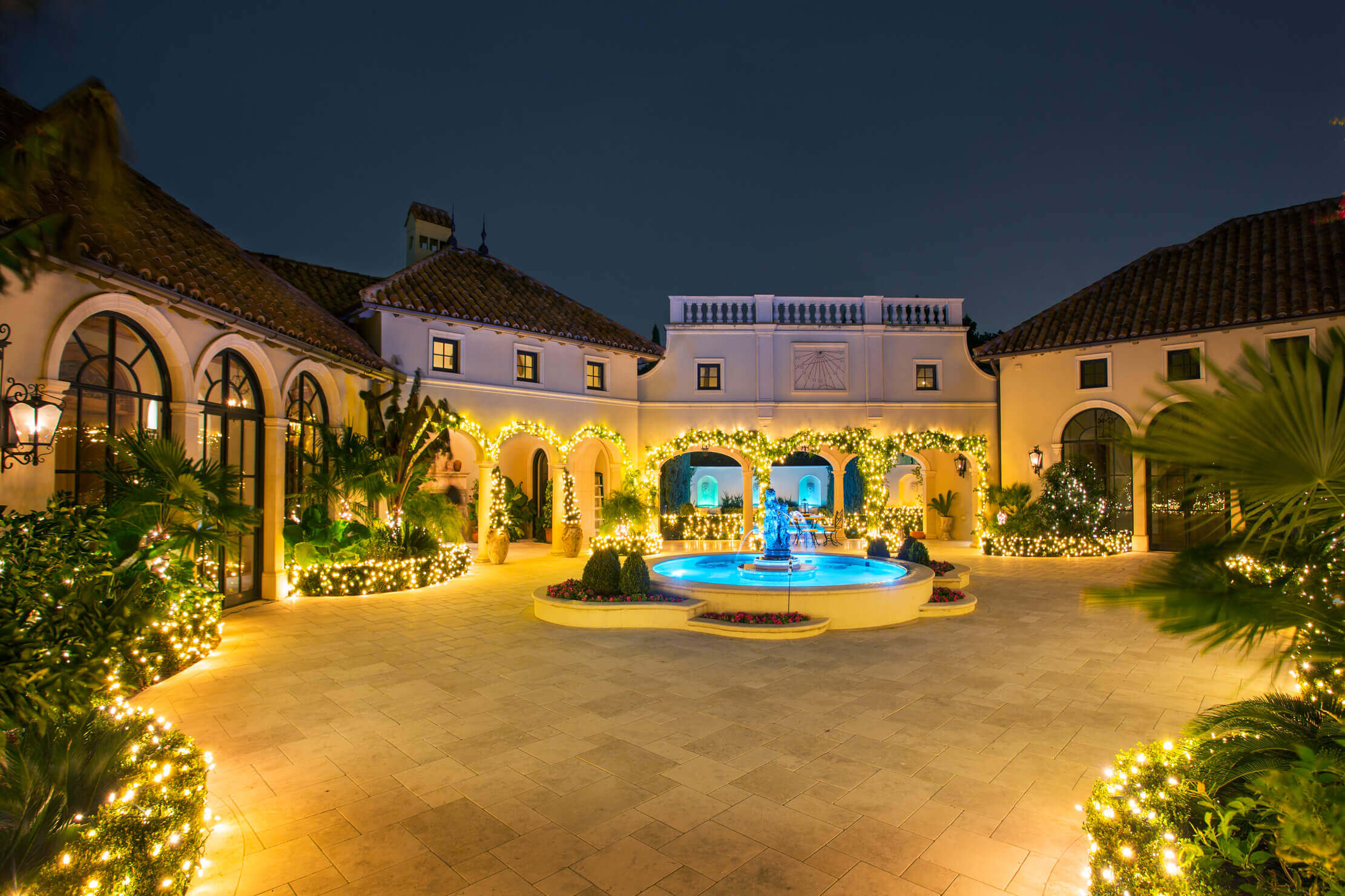 Elegant courtyard at night featuring a fountain, surrounded by greenery and twinkling lights on buildings and paths.
