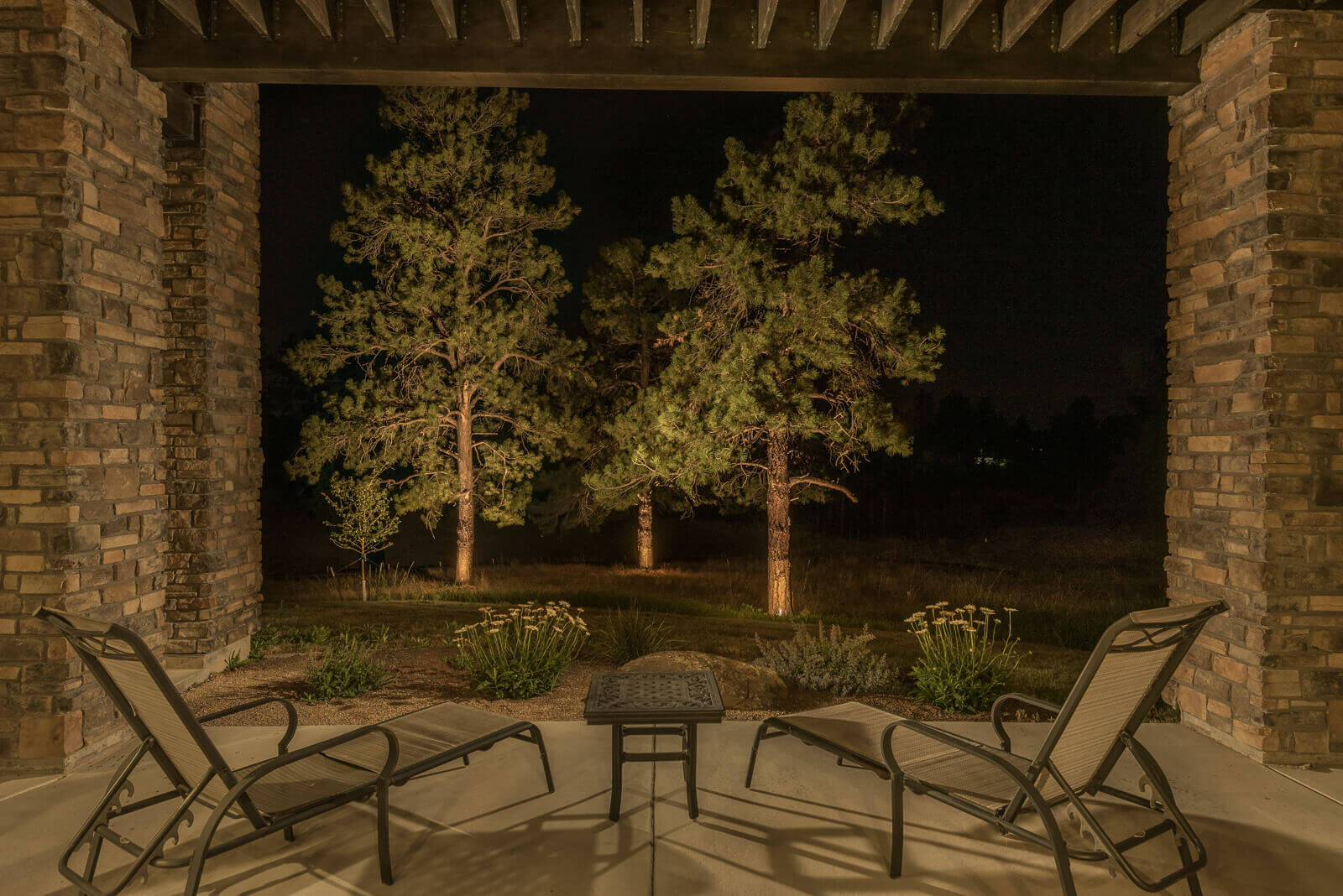 Night view of a patio with two lounge chairs and a table, surrounded by lit pine trees and landscaping.